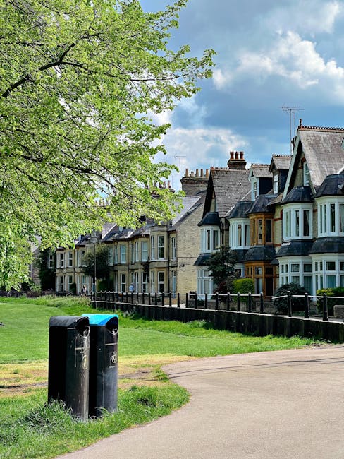 A row of residential houses with traditional architecture, featuring bay windows and slate roofs, situated along a paved sidewalk. In the foreground, two black cylindrical waste bins are placed beside